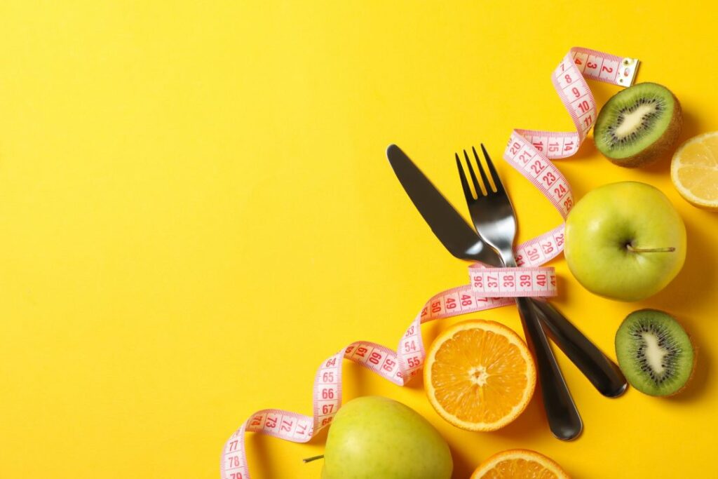 Healthy meal planning concept with fresh fruits, a blue measuring tape, and silver cutlery on a vibrant yellow background.